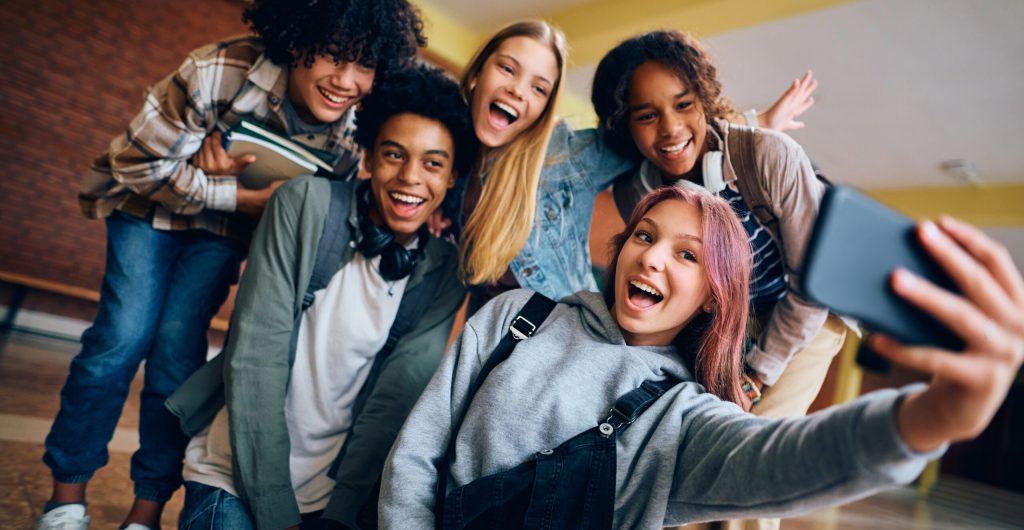 Group of happy high school students having fun while taking selfie with cell phone in hallway.